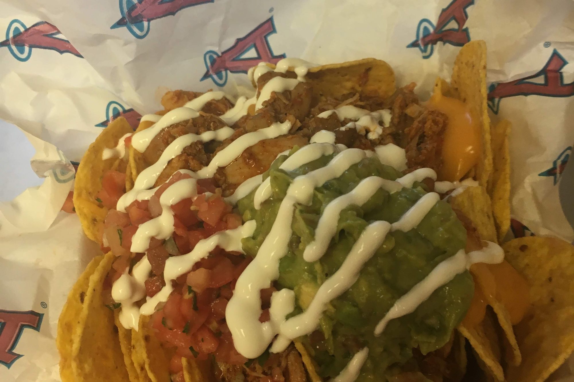 A tray of loaded nachos at Angel Stadium, chips under shredded chicken, nacho cheese, pico de gallo, guacamole and a drizzle of sour cream, sitting on paper printed with the Angels halo logo
