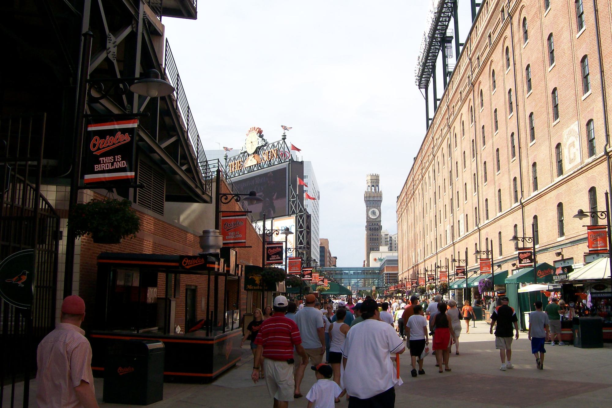 Fans walking the Eutaw Street concession row at Camden Yards, stands on both sides, the brick B&O warehouse to the right and the Bromo Seltzer tower in the distance
