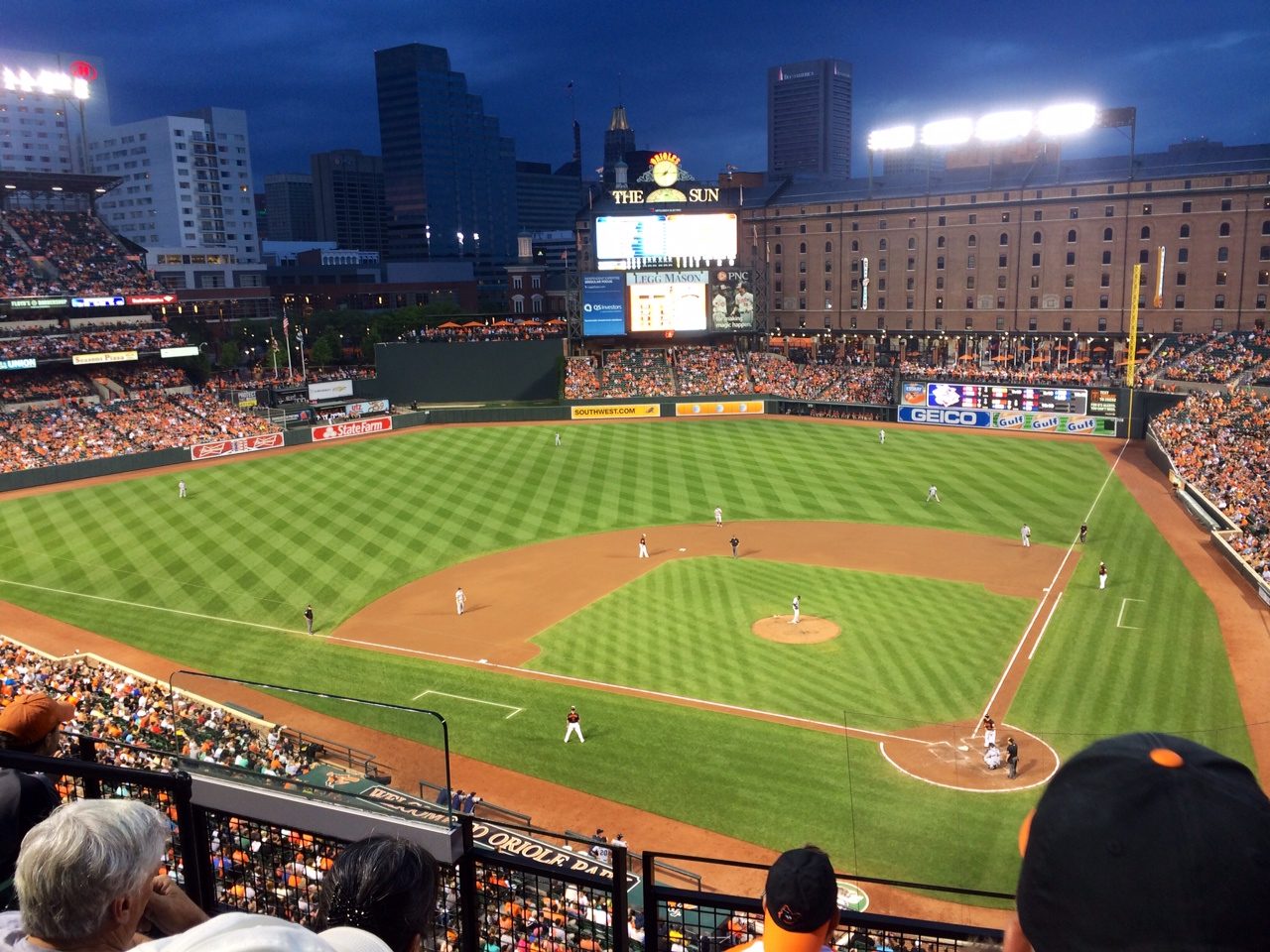 Dusk settling over Camden Yards during a summer night game, the warehouse and the Baltimore skyline behind the outfield