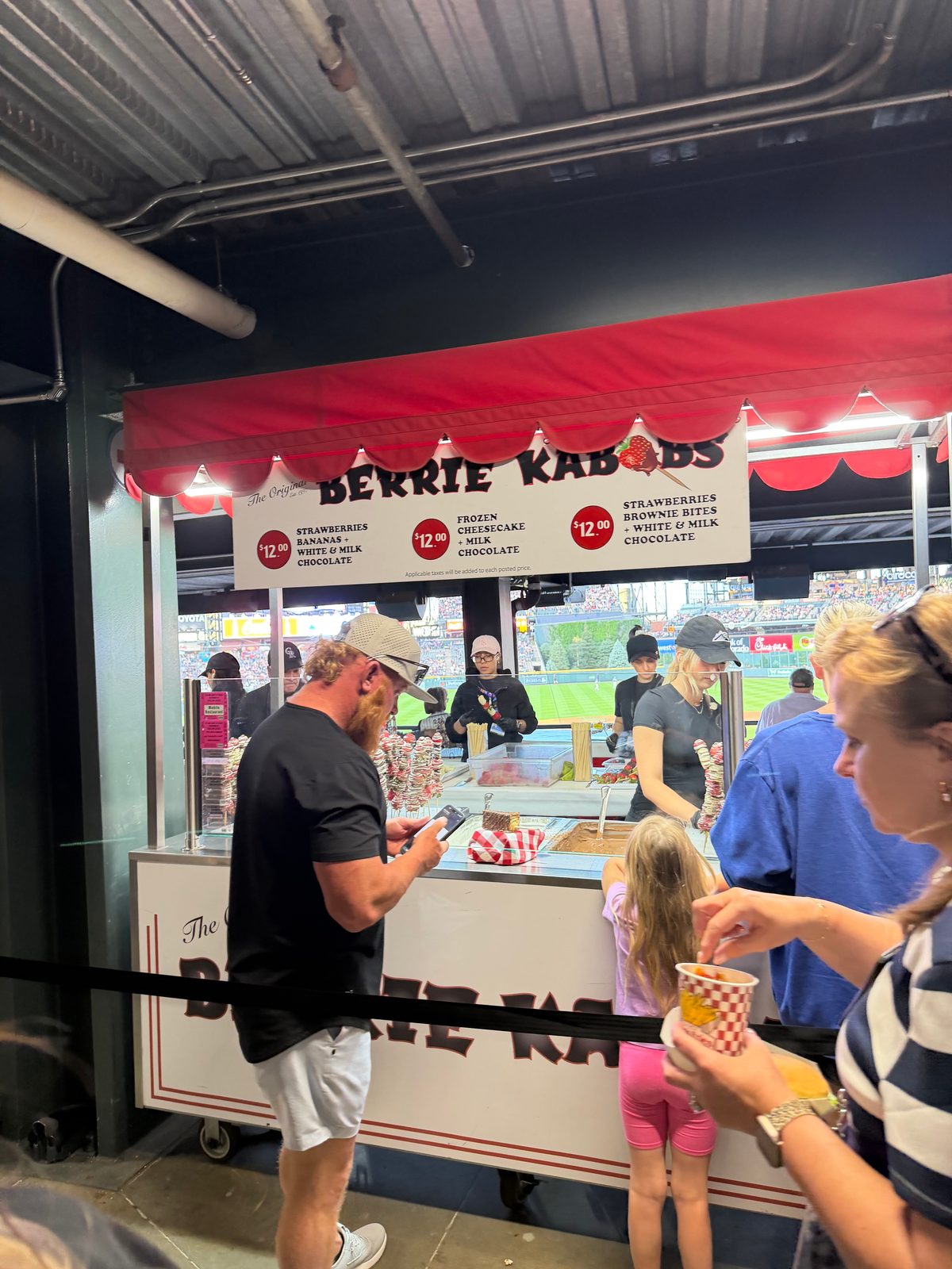 The Berrie Kabobs stand at Coors Field under its red scalloped awning, the menu board and racks of chocolate-covered kabobs visible at the counter with the field beyond