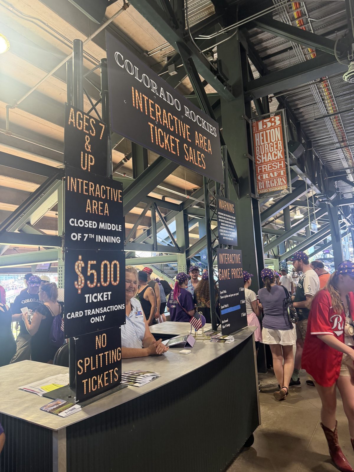 The Colorado Rockies Interactive Area ticket sales window at Coors Field, lit marquee signage reading Interactive Area Ticket Sales, Ages 7 and Up, and Interactive Area Closed Middle of 7th Inning, with a staff member at the counter and fans on the concourse