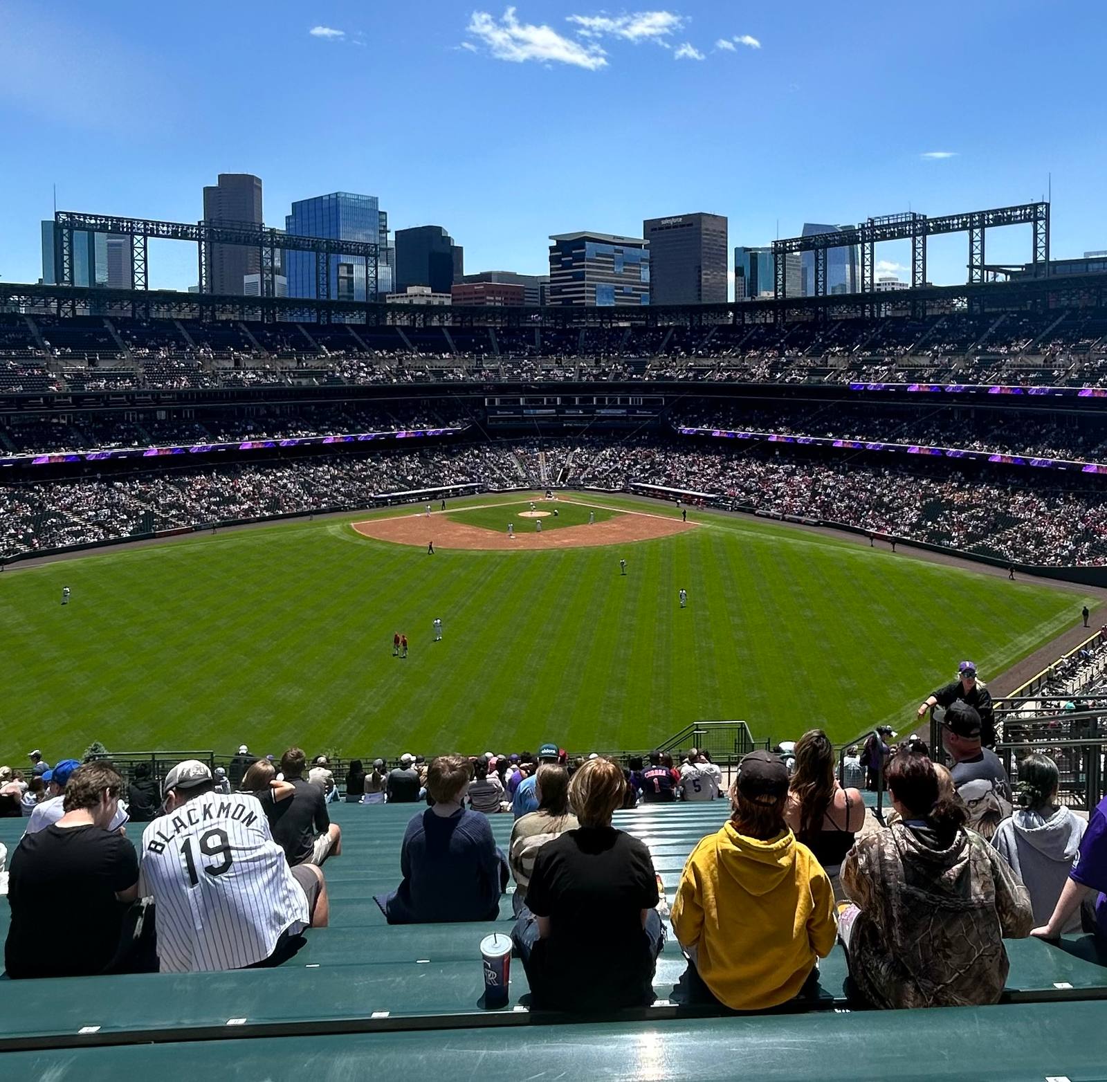 The view from the Rockpile bleachers in dead center field at Coors Field, looking back across the outfield at home plate with the downtown Denver skyline rising beyond the grandstand