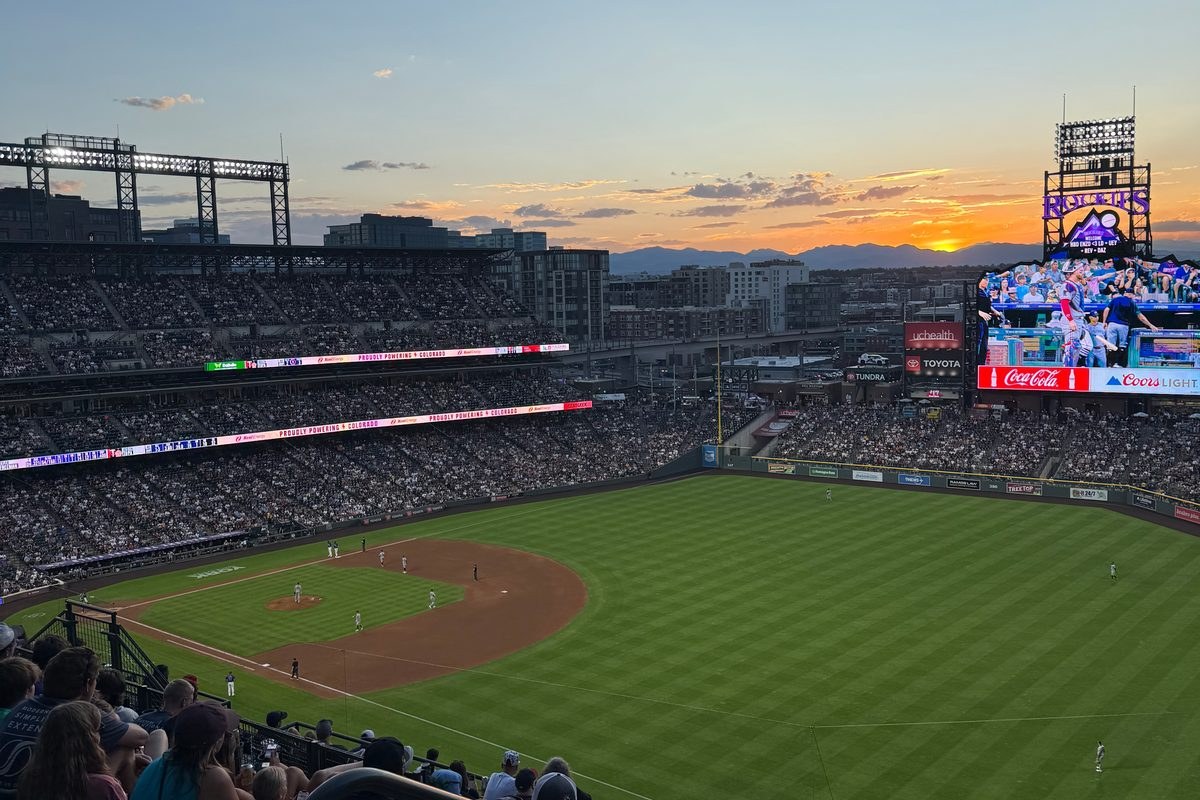 Sunset over Coors Field during a game, the outfield and videoboard under a pink and orange sky