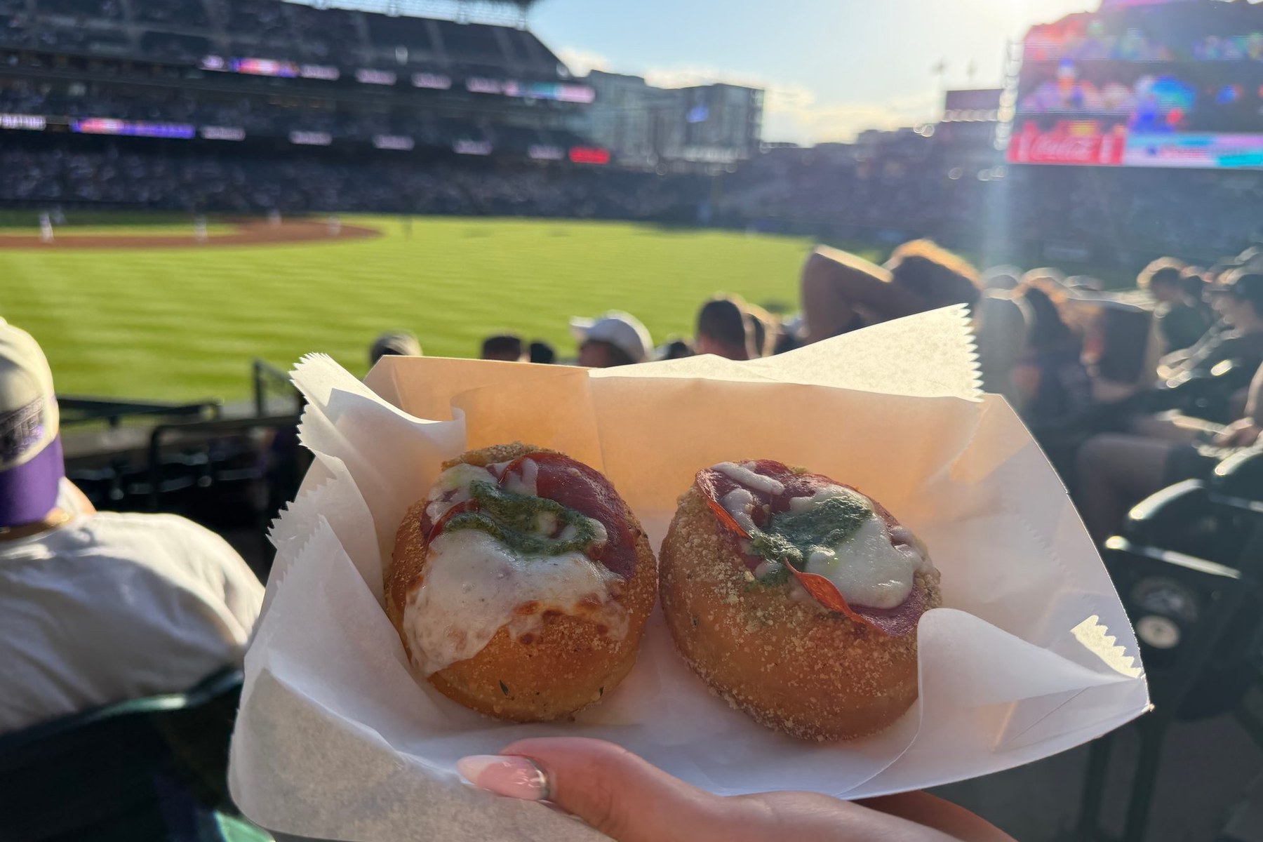 Two pizza donuts in a paper tray held up in the stands at Coors Field, with the outfield grass and the crowd behind