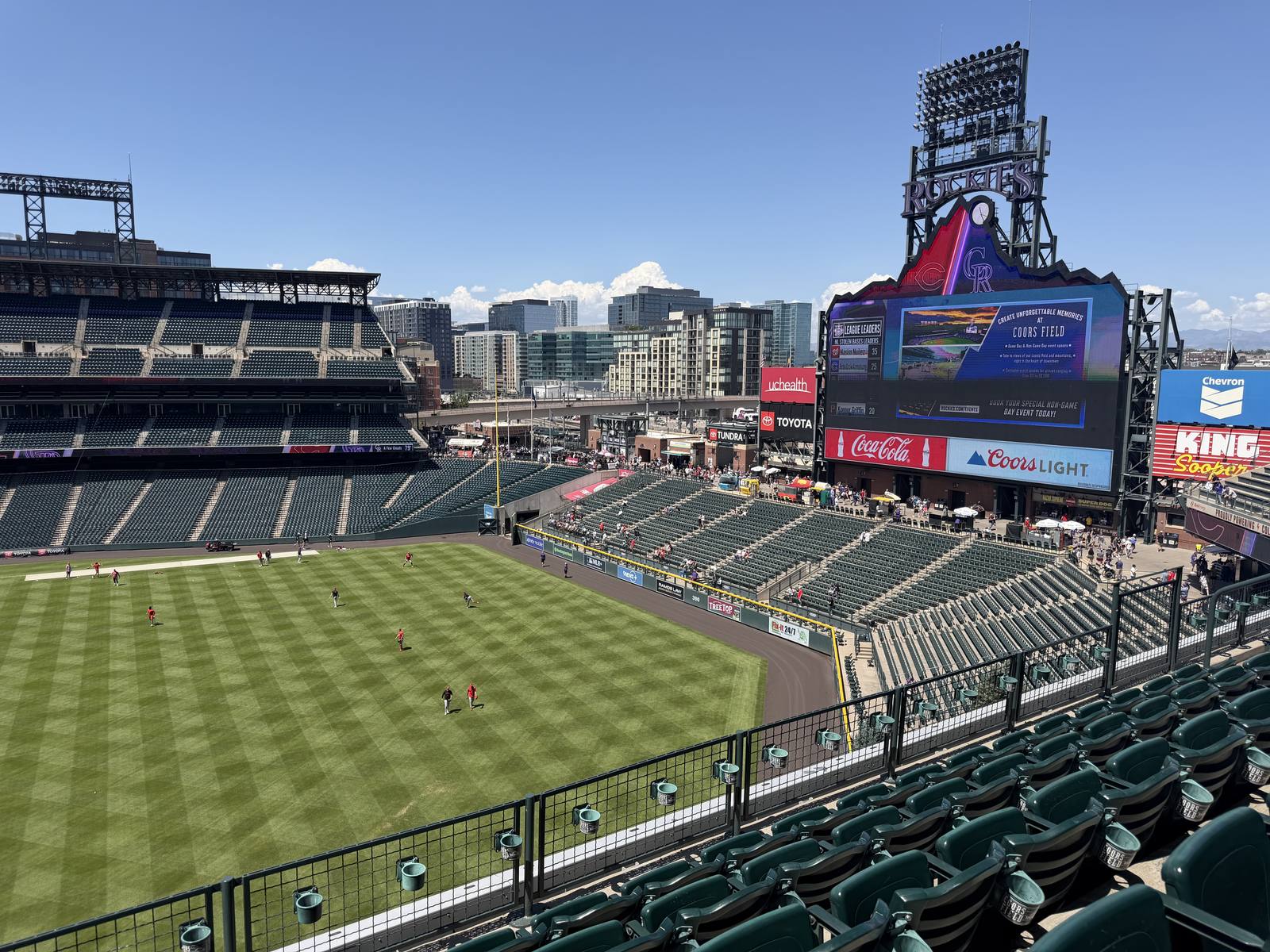 The mostly empty seating bowl at Coors Field before a day game, players warming up on the outfield grass with the scoreboard and downtown Denver skyline beyond under a clear blue sky