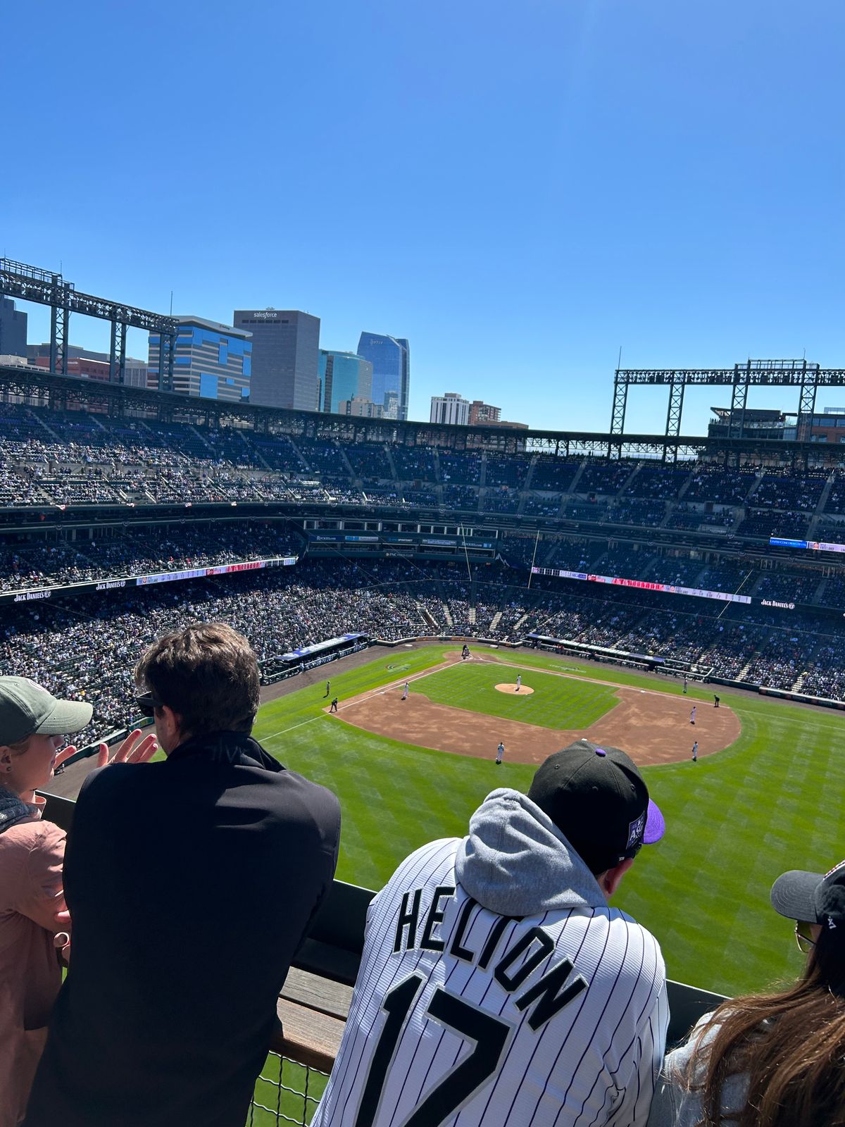 The view from the top of the stadium at Coors Field, the full seating bowl and field below and the downtown Denver skyline beyond, a fan in a Helton jersey at the rail in the foreground