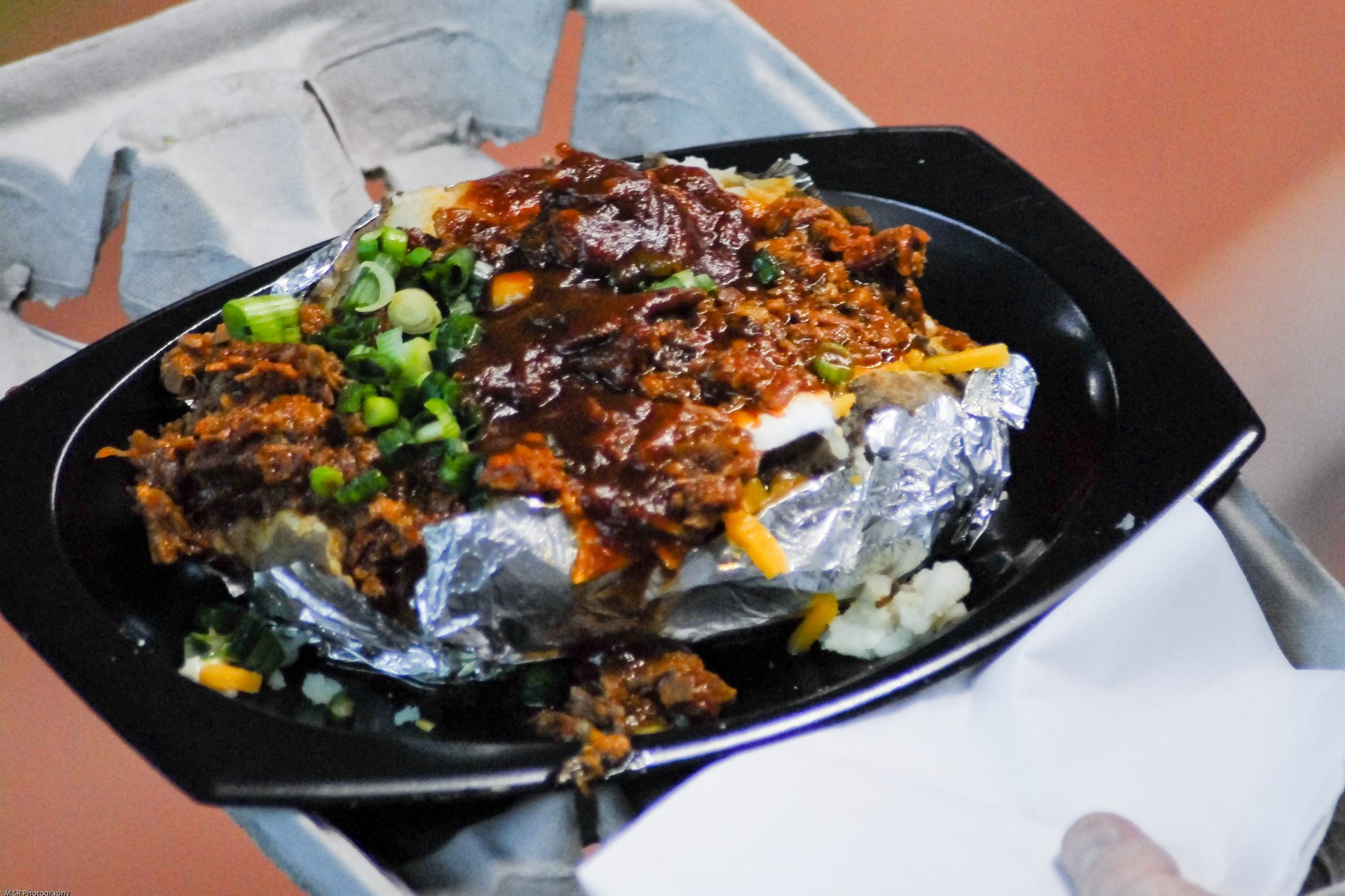 A barbecue-loaded stuffed baked potato in foil on a black tray, topped with brisket, barbecue sauce, cheddar and green onion, held on a lap at a Houston Astros game