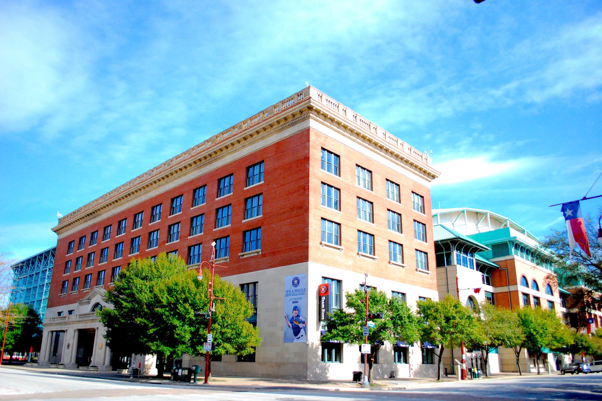 The red brick Union Station head house on the corner of Crawford Street and Texas Avenue, with the glass wall and teal roof of Daikin Park rising behind it and a Texas flag hanging over the sidewalk