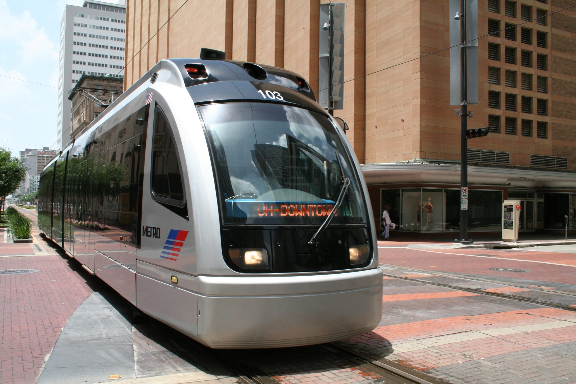 A METRORail light rail car stopped on a brick-paved downtown Houston street, its headsign reading UH-Downtown, with office towers behind it