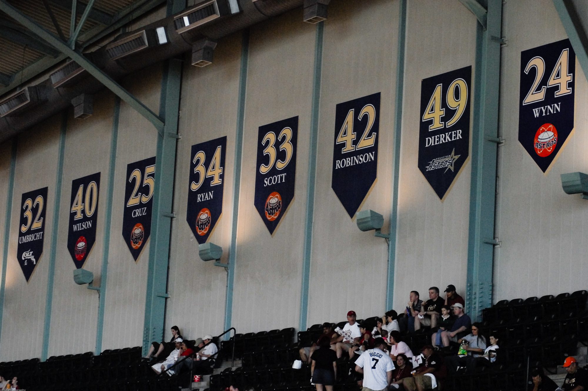 Retired-number banners hanging on the wall at Daikin Park, among them 32 Umbricht carrying the Colt .45s logo, 34 Ryan, 33 Scott, 42 Robinson, 49 Dierker and 24 Wynn, with fans in the seats below