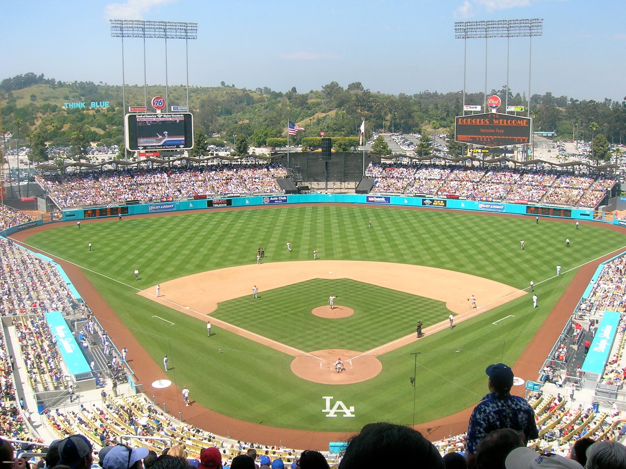 A day game at Dodger Stadium seen from high behind home plate, with both outfield pavilions, the two scoreboards, and the wooded hills beyond the outfield