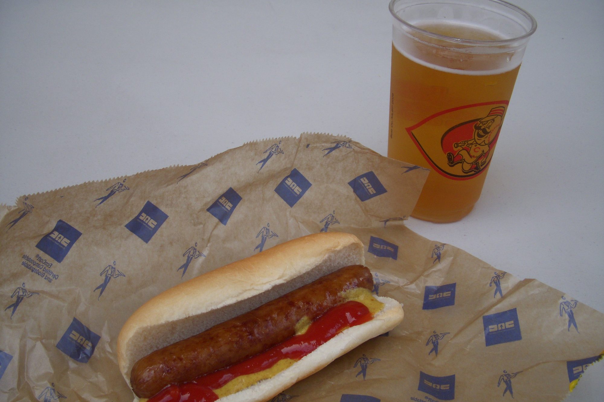 A Big Red Smokey at Great American Ball Park, a long grilled sausage in a bun with ketchup and mustard on its printed paper wrapper, next to a clear cup of beer carrying the Reds' Mr. Redlegs logo