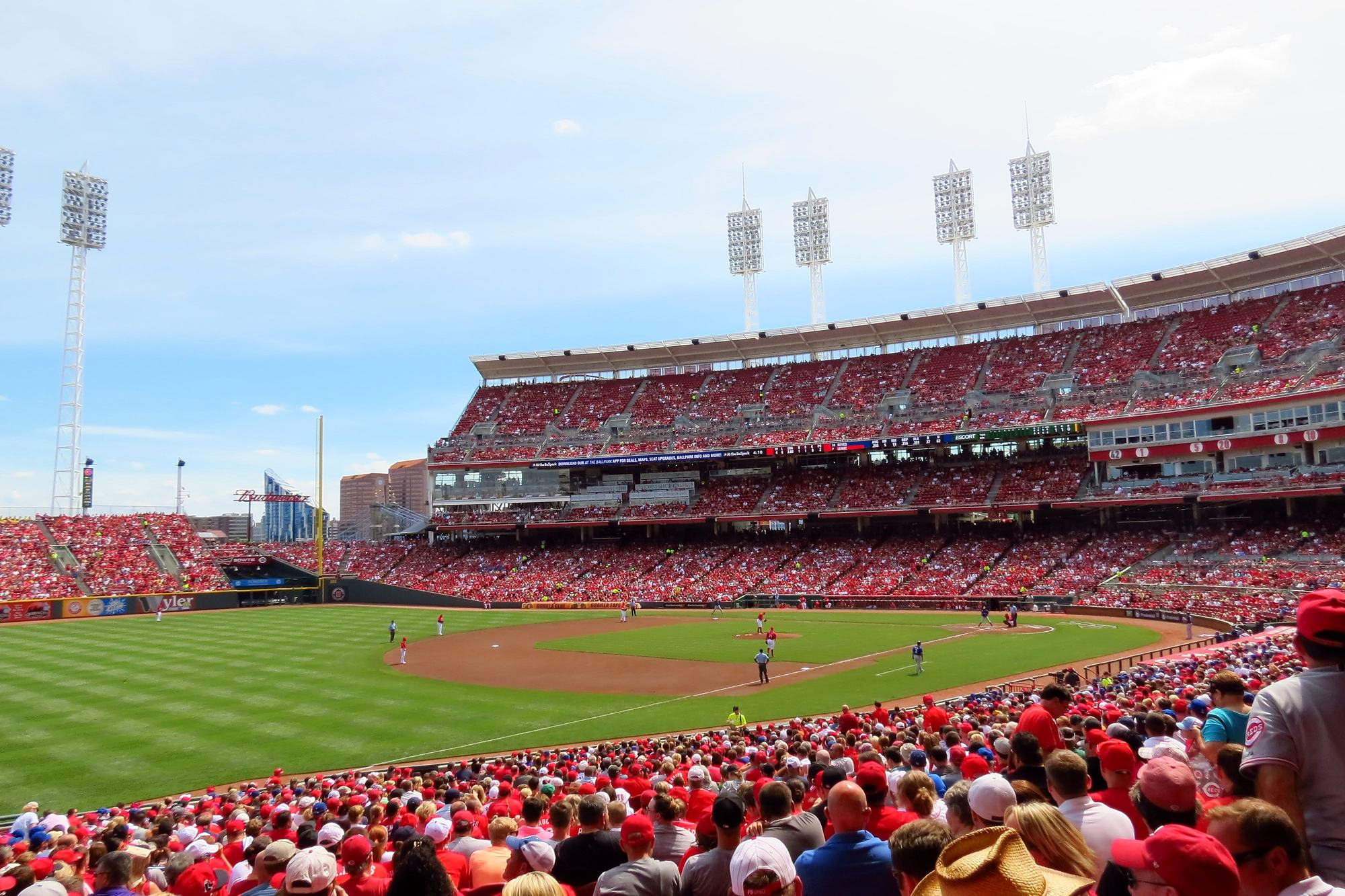 A full house at Great American Ball Park on a sunny afternoon, seen from a seat down the line, fans packing the lower bowl in the foreground with the infield, the tiered grandstand, and the light towers beyond