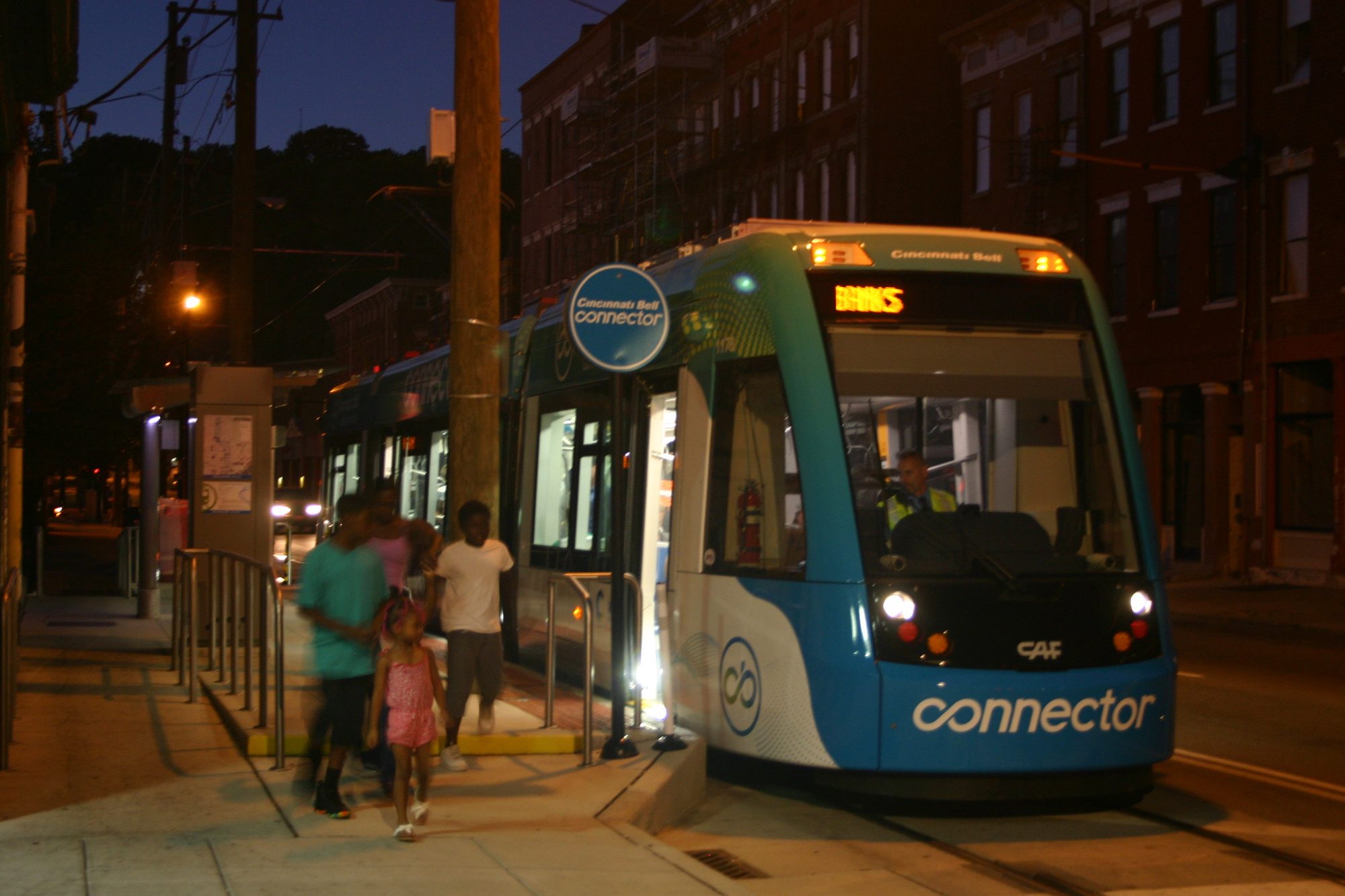 A Connector streetcar stopped at a station at dusk, a family stepping off onto the platform, the destination sign reading BANKS and brick storefronts lining the street