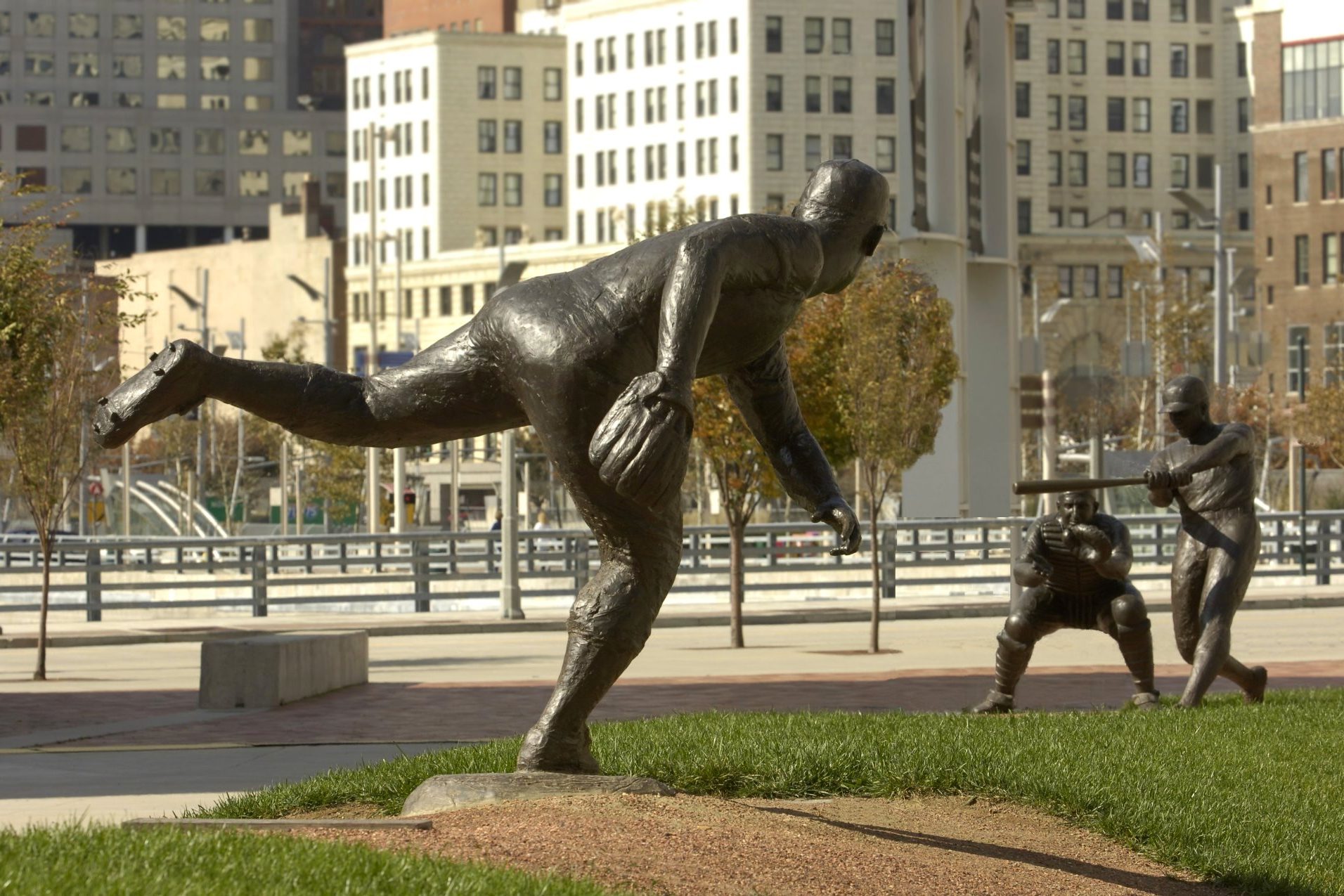 The bronze Reds legends on Crosley Terrace outside Great American Ball Park, a pitcher mid-delivery in the foreground with a catcher crouched and a batter set behind him, downtown Cincinnati buildings across the street