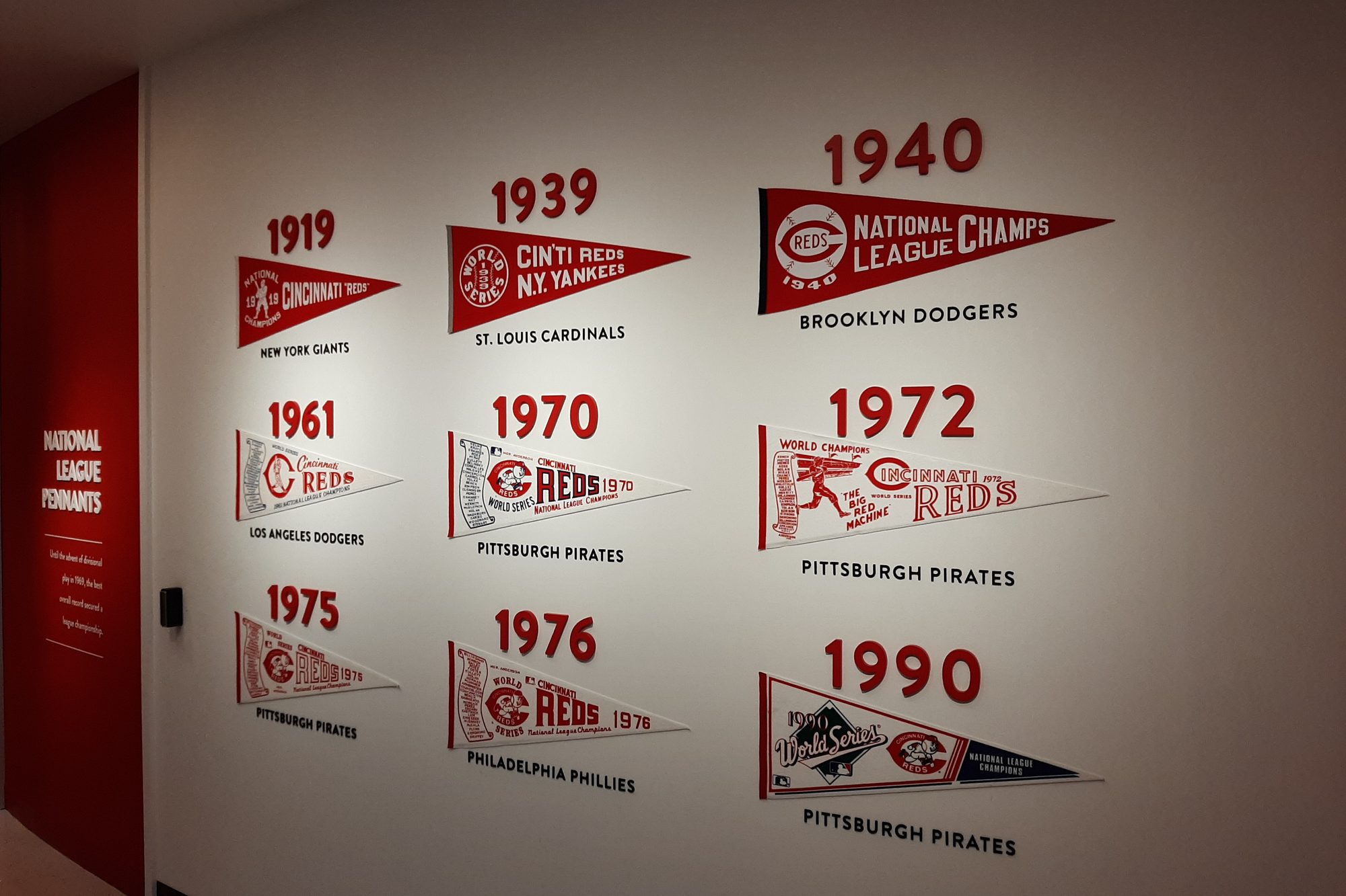 The National League pennants display inside the Reds Hall of Fame and Museum at Great American Ball Park, nine pennants from 1919 through 1990 mounted in rows on a white wall with the year above each one