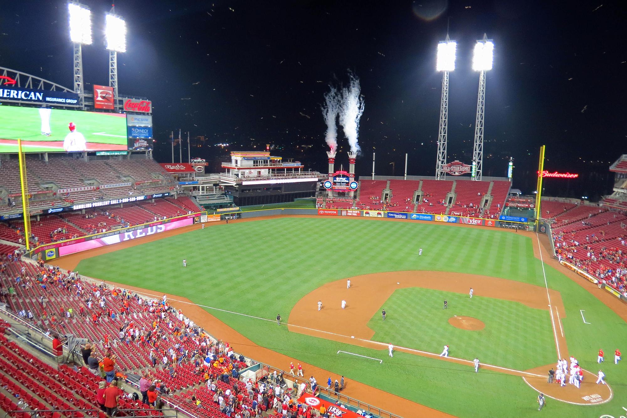 A night game at Great American Ball Park seen from the upper deck, the Power Stacks firing above right-center field under the lights, players gathered on the infield as the crowd empties out