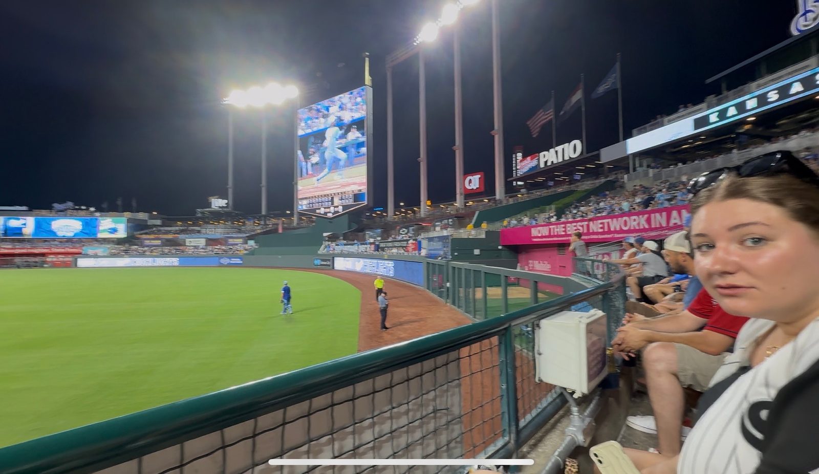 A night view from the outfield patio at Kauffman Stadium looking in toward the lit crown scoreboard, with the bowl glowing under the lights and fans along the rail