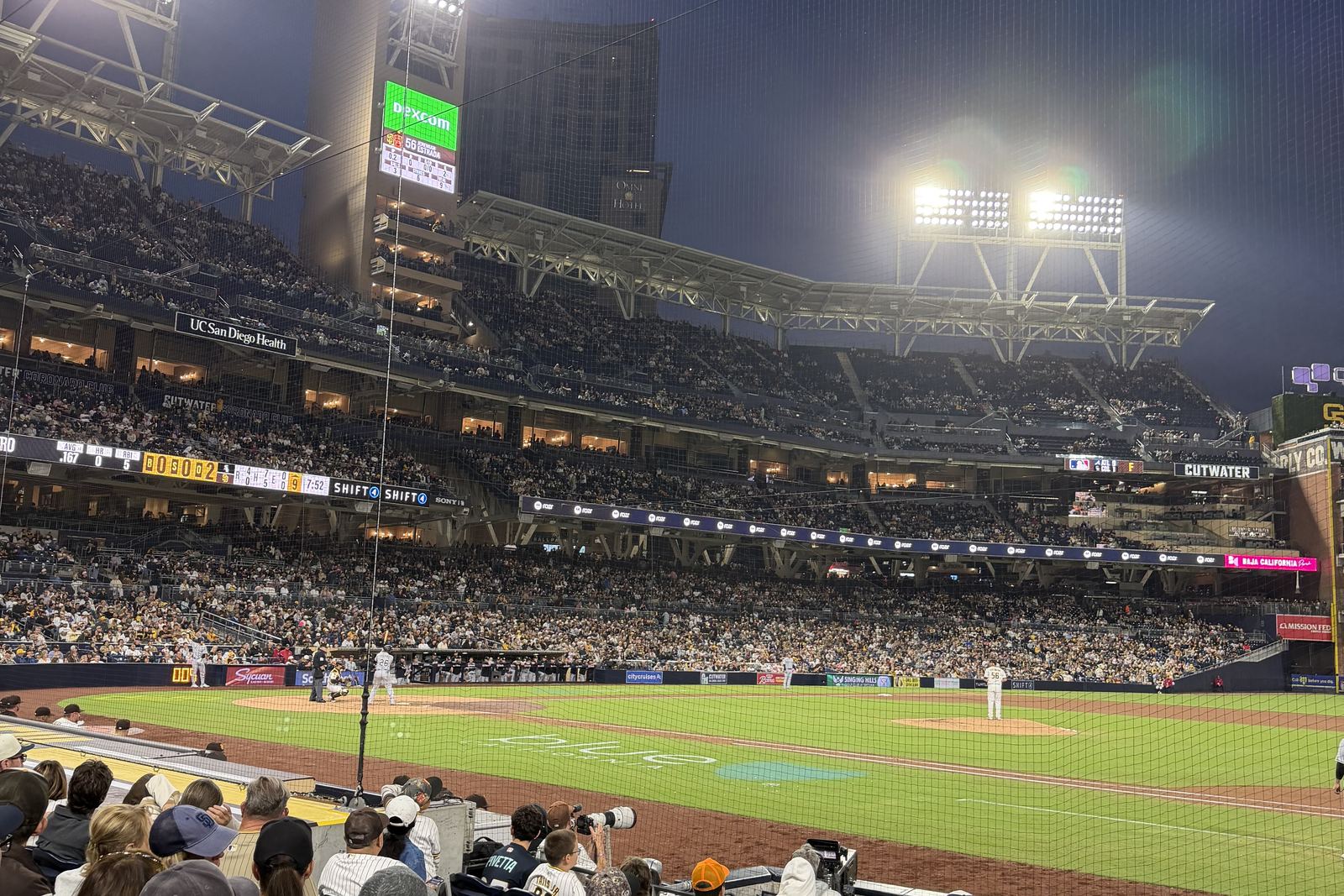 A packed night game at Petco Park seen from the lower bowl behind home plate, the light standards on and the downtown San Diego towers behind the outfield
