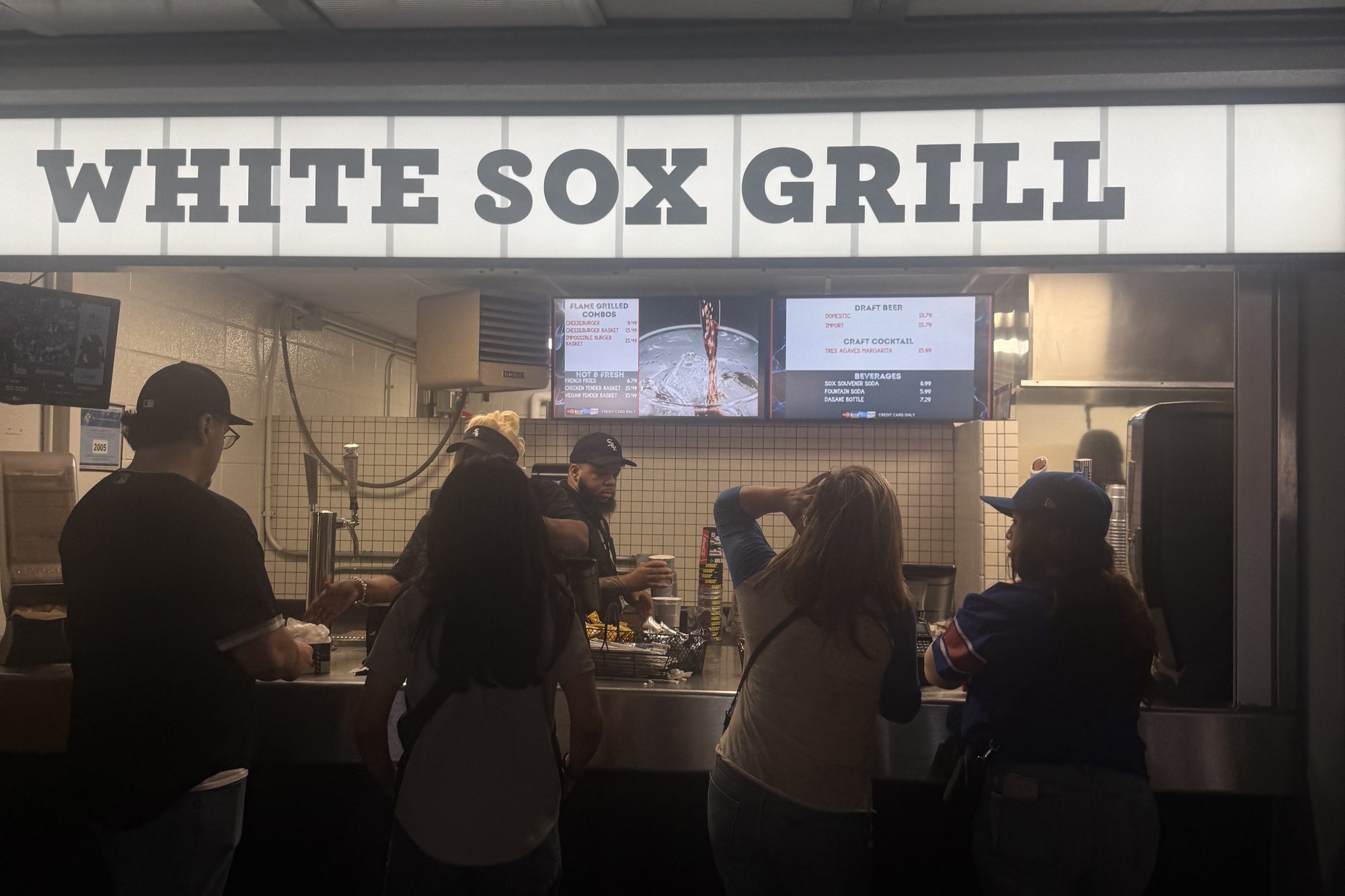 Fans lined up at the White Sox Grill counter on the concourse at Rate Field, the lit stand sign above them and the menu boards showing burgers, baskets and draft beer