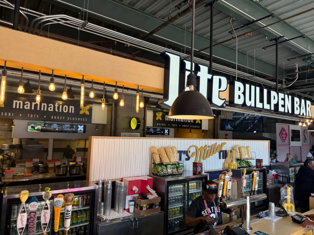 The Miller Lite Bullpen Bar counter at T-Mobile Park with its beer taps and a bartender, and the Marination stand sign just to the left