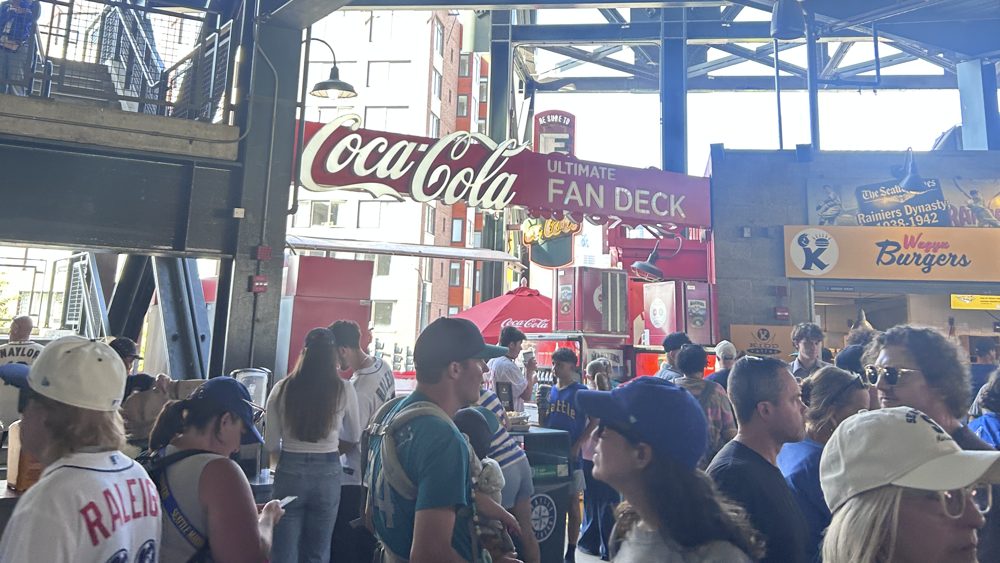 A crowd on the concourse at T-Mobile Park below the Coca-Cola Ultimate Fan Deck sign, with a Wagyu Burgers stand to the right