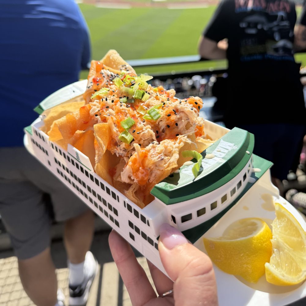 Crab nachos on fried wonton chips with green onion, black sesame, and lemon wedges, served in a white and green ferry-shaped souvenir tray held up over the field at T-Mobile Park