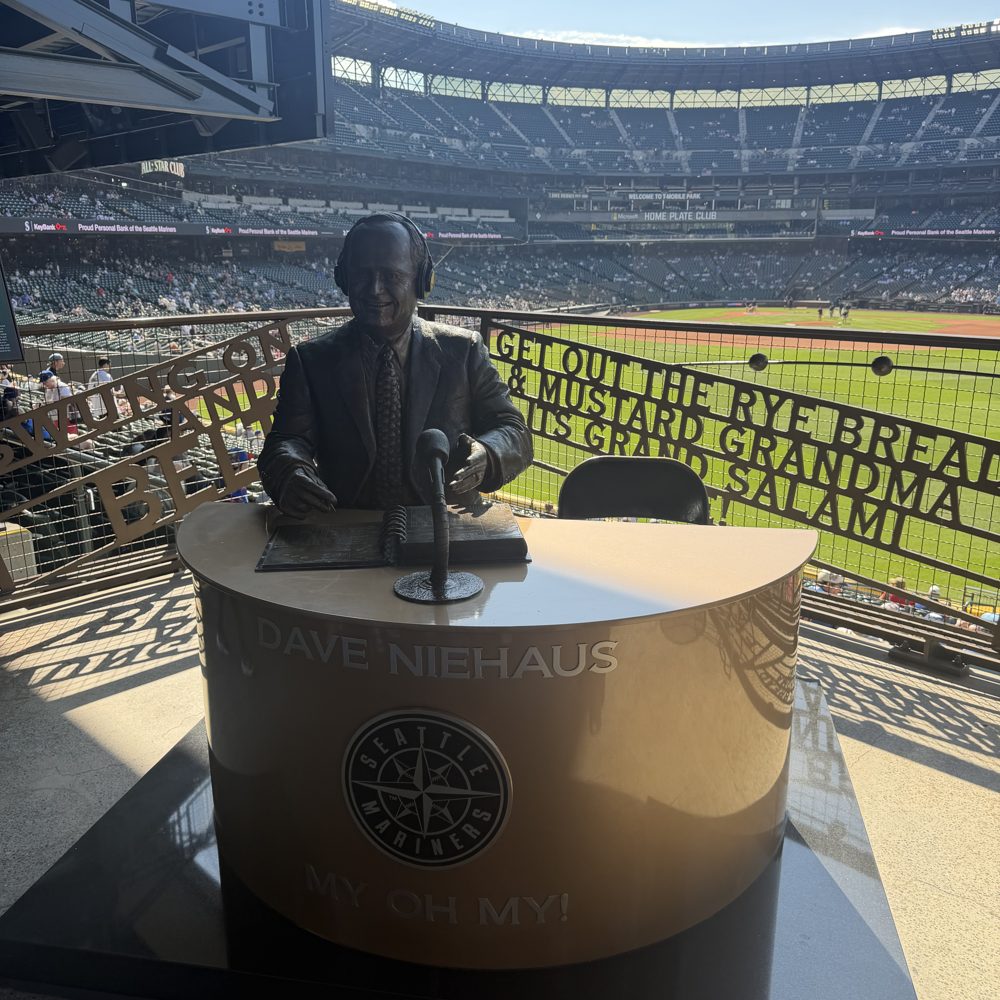 The bronze statue of broadcaster Dave Niehaus at a broadcast desk inside T-Mobile Park, with the field behind and his calls cut into the railing