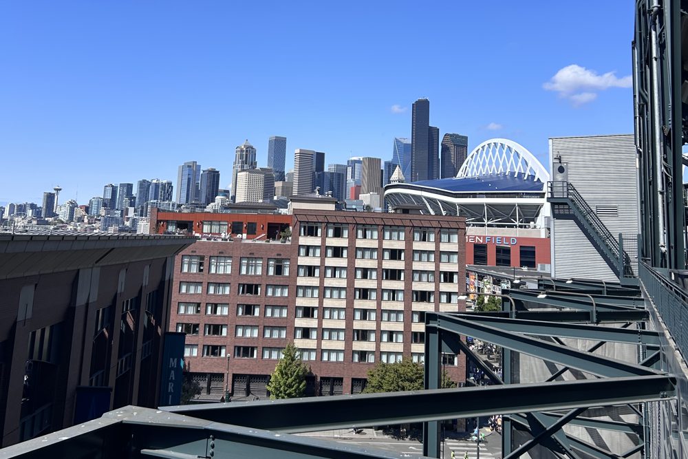 The downtown Seattle skyline and the roof of Lumen Field seen from the upper deck of T-Mobile Park, framed by the stadium's steel