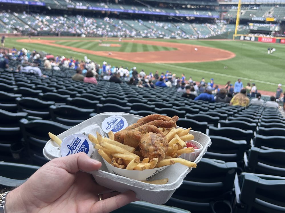 A tray of Ivar's fish and chips with tartar sauce cups, held over the lower-level seats at T-Mobile Park with the field behind