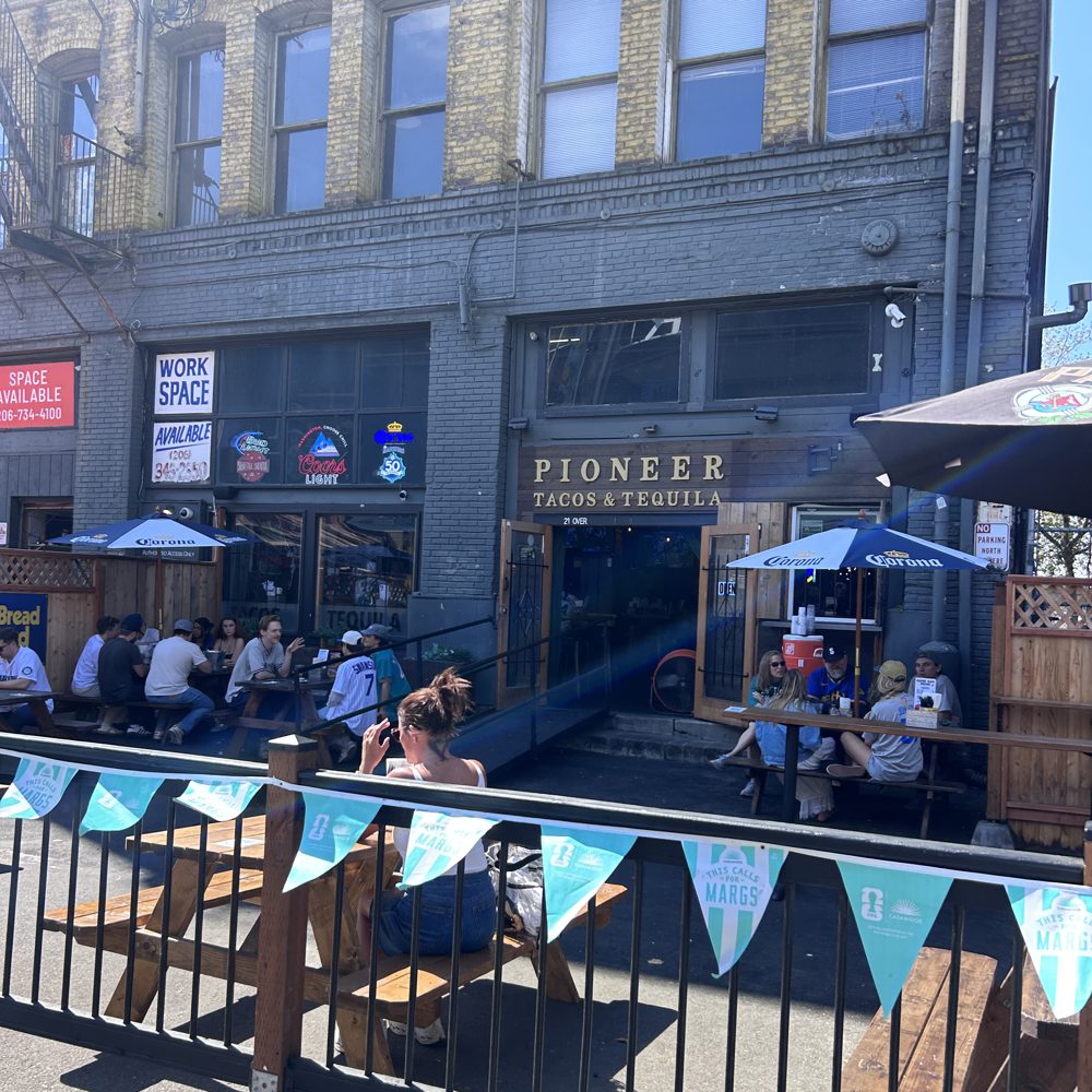 The patio outside Pioneer Tacos and Tequila in Seattle, with fans at picnic tables behind a railing hung with pennant bunting on a sunny day