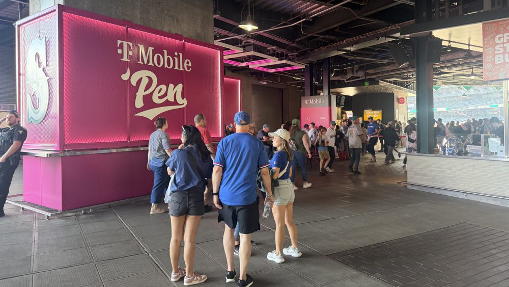 The pink T-Mobile 'Pen sign on a kiosk under the concourse deck at T-Mobile Park, with fans walking past toward the outfield