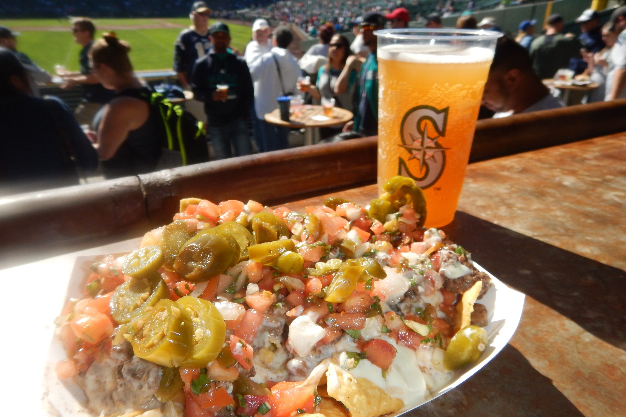Loaded nachos and a Mariners-branded beer cup on a wooden drink rail at T-Mobile Park, with standing-room tables and the field beyond