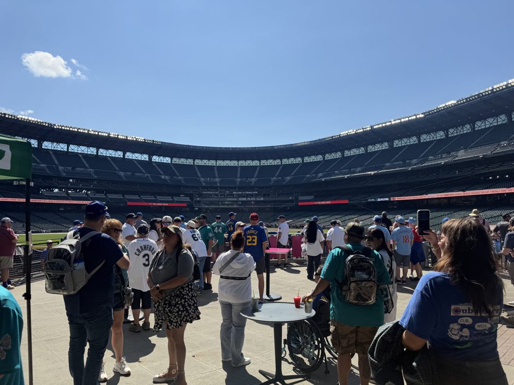 Fans standing around drink tables in an outfield standing-room area at T-Mobile Park, with the grandstand across the field