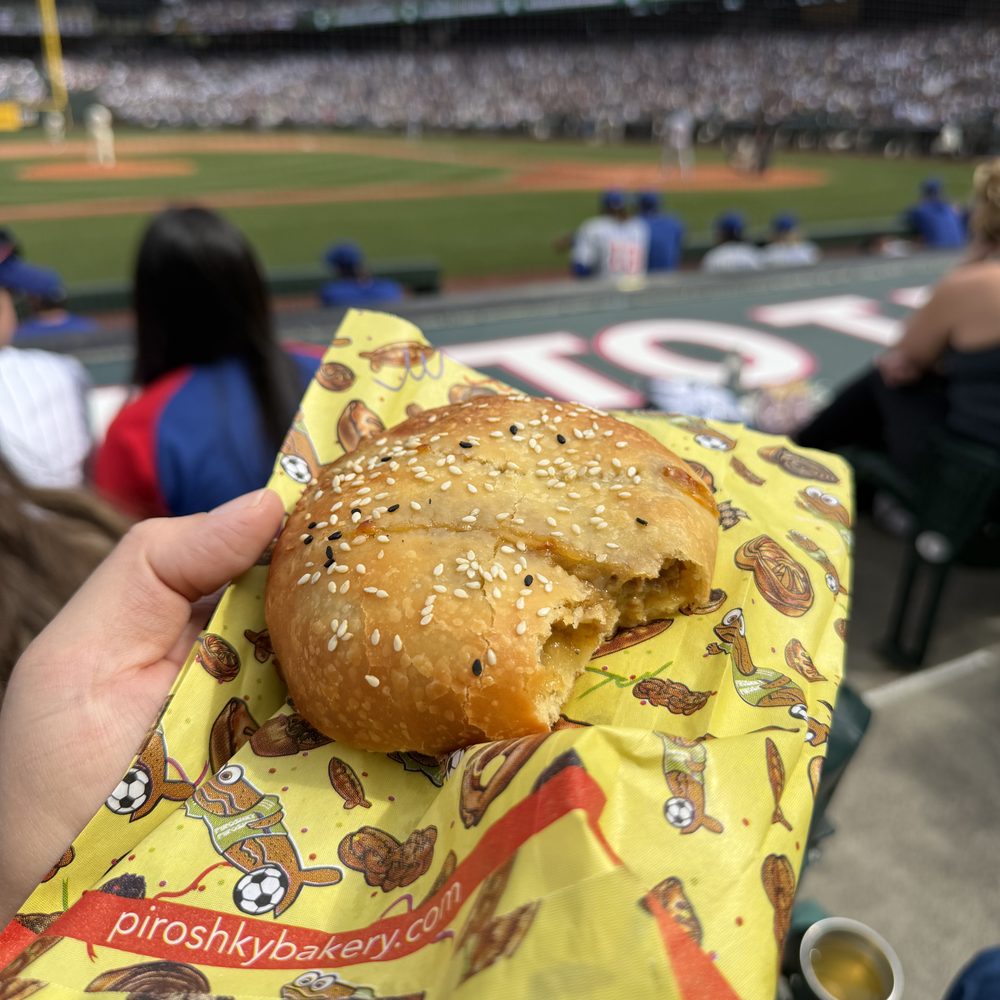 A sesame-topped piroshky with a bite taken out of it, held on a yellow Piroshky Bakery wrapper above the lower-level seats at T-Mobile Park, with the field behind