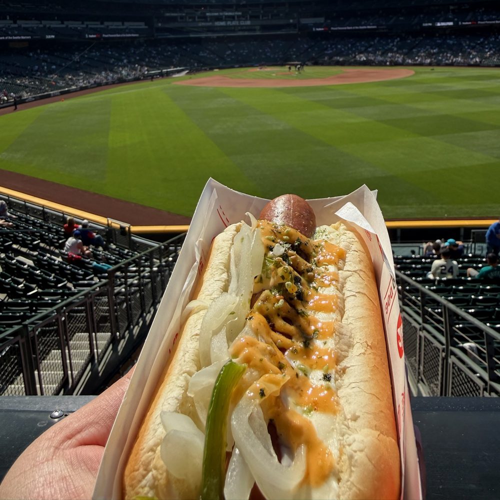 The Sumo Dog Seattle Dog, a hot dog topped with onions, a green pepper, furikake, and an orange sauce, held over the upper-deck rail at T-Mobile Park
