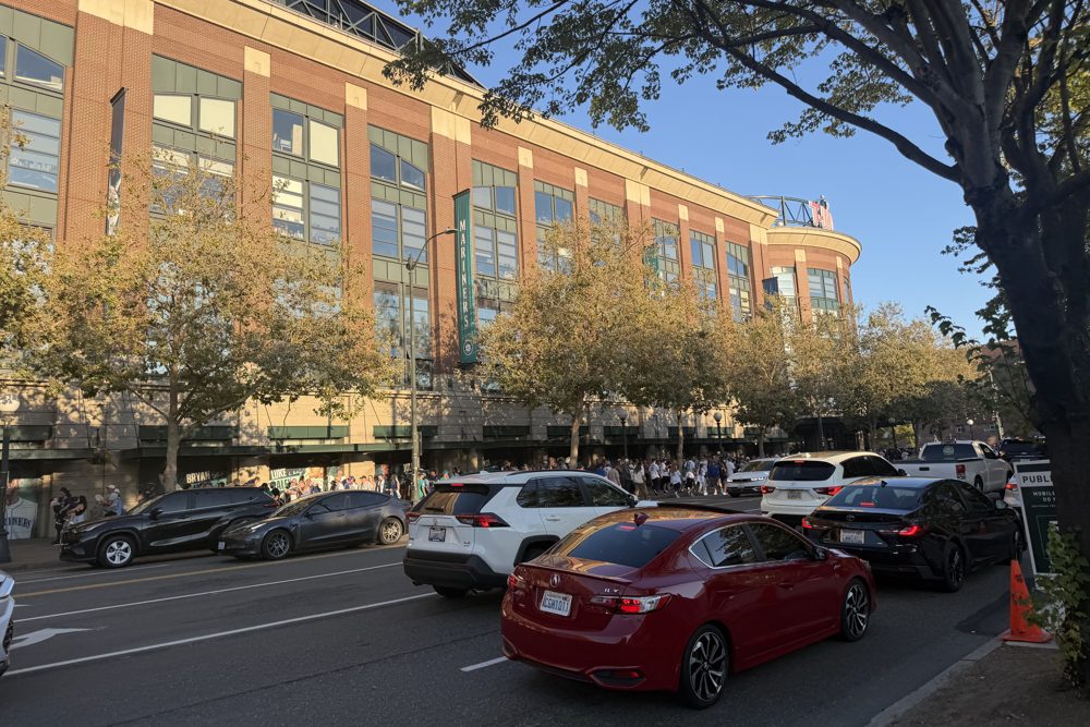 Cars lined up on the street along the brick side of T-Mobile Park, with trees and a Mariners banner on the building