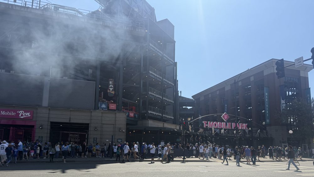 Fans crossing the street toward T-Mobile Park on a sunny afternoon, with grill smoke drifting across the front of the stadium