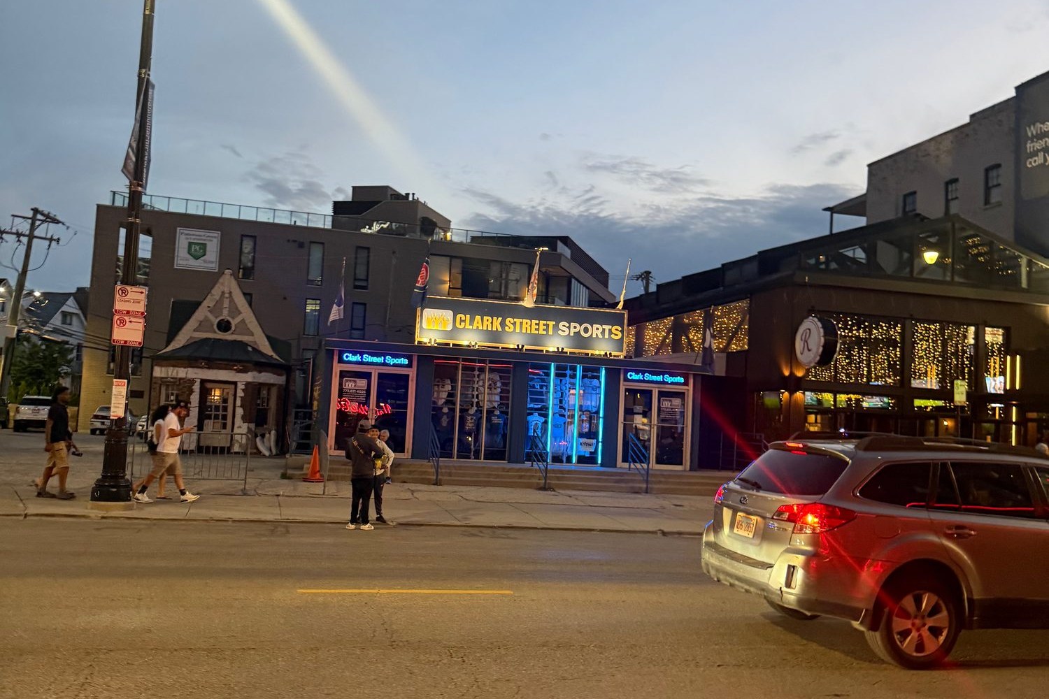 The Clark Street corner in Wrigleyville at dusk, bars and storefronts lit up across from Wrigley Field