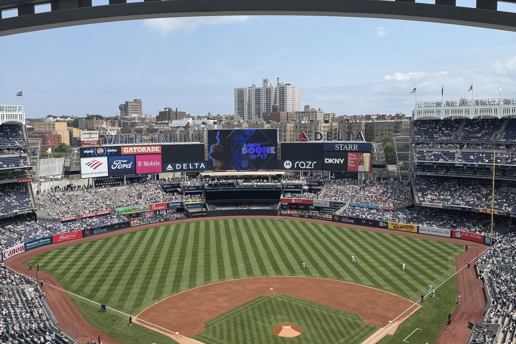 The open-air bowl at Yankee Stadium from the upper Grandstand behind home plate, the white frieze running along the far roofline, the videoboard and the Bronx apartment blocks beyond the outfield