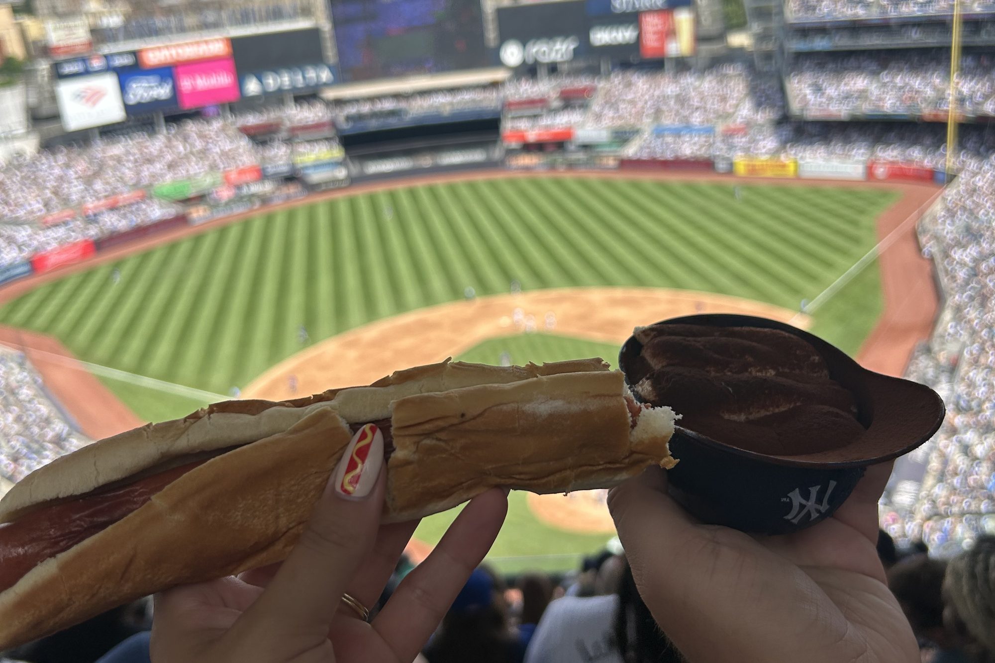 A hot dog and a chocolate soft-serve sundae in a Yankees batting-helmet cup, held up over the packed bowl at Yankee Stadium, the infield and the outfield seats behind them